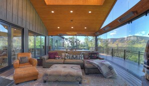 A spacious living area with a large sofa, ottoman, and armchair, a wooden ceiling, and a view of a mountainous landscape through large windows.