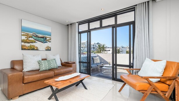 A modern living room with a brown leather sofa, a wooden coffee table, and an orange chair. There is a large window with curtains, a painting on the wall, and a view of the outside. - Eden On The Bay 104 (Cape Town)