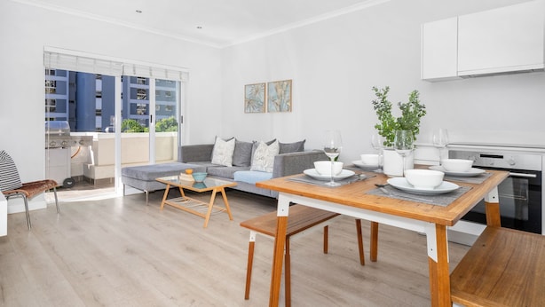 A modern kitchen with white cabinets, a dining table with benches, and a large window overlooking a balcony. - Zanzibar Mansions (Cape Town)