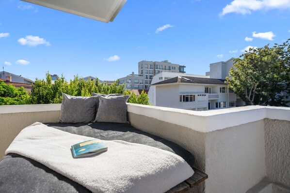 Un balcón con sofá, almohadas y un libro. El balcón da a una zona residencial con edificios modernos y zonas verdes.