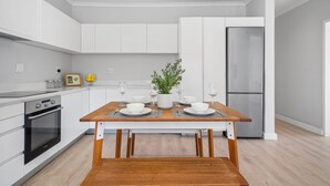 A modern kitchen with white cabinetry, a wooden dining table, and a built-in oven. - Zanzibar Mansions (Cape Town)