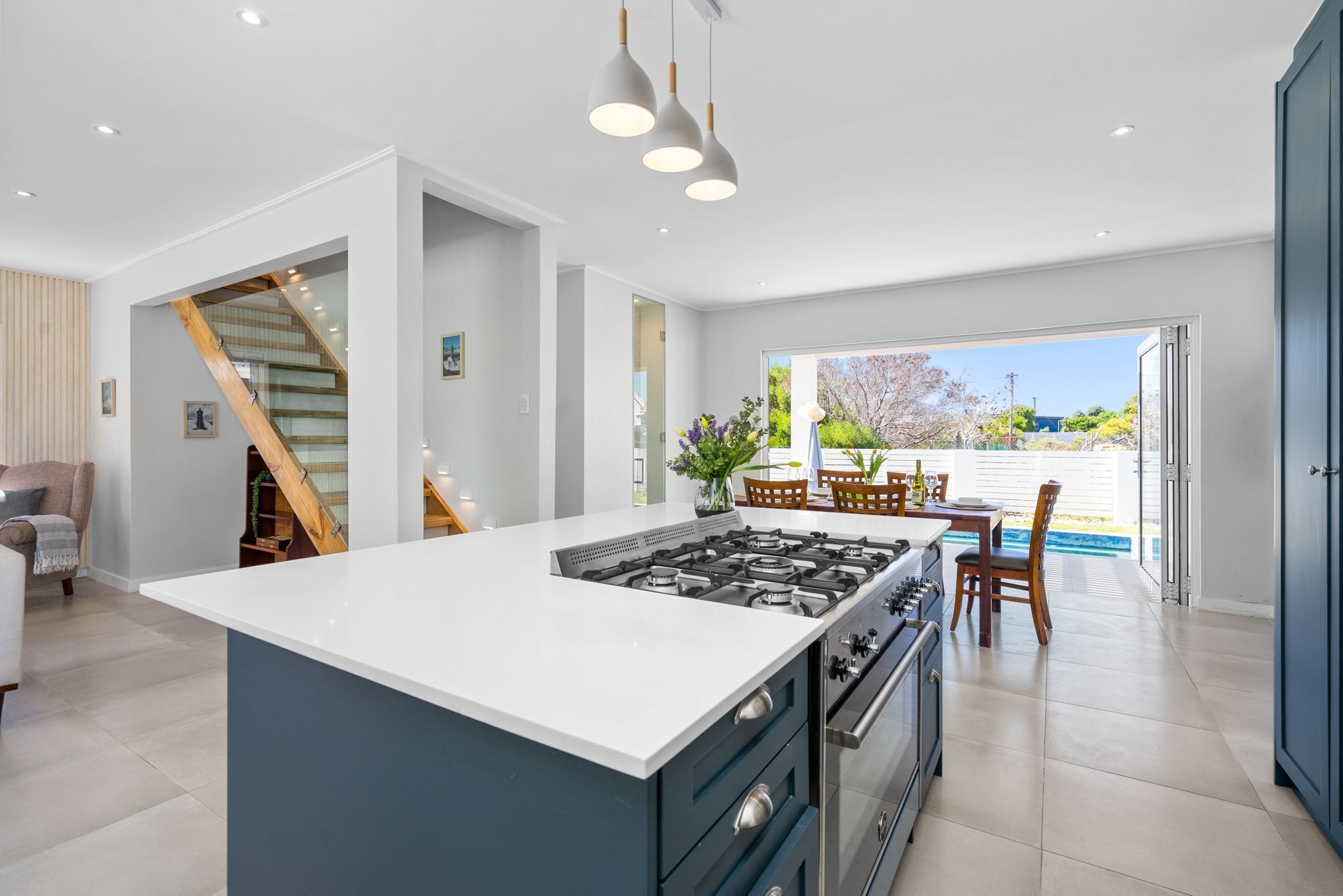A modern kitchen with a central island, a dining area with wooden chairs, and a staircase leading to an upper floor.