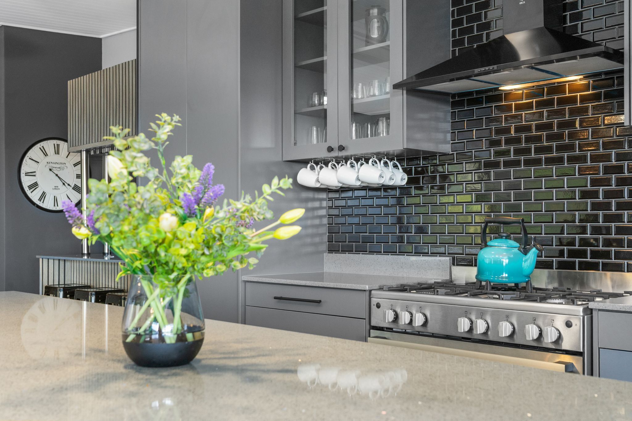 Une cuisine moderne avec des meubles gris, un dosseret en carreaux noirs, une cuisinière en acier inoxydable et une bouilloire turquoise.