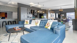 A modern living room with a blue sectional sofa, a coffee table, and a kitchen area with a bar. - Red River Valley (Rooiels)