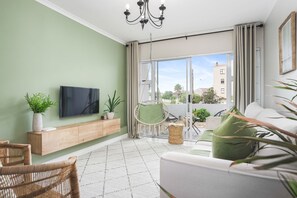 A modern living room with a flat-screen TV, wooden console, wicker chairs, a green sofa, and a balcony with a view of buildings and greenery. - The Sands BG3 (Cape Town)
