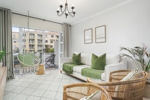 A modern living room with a white sofa, green cushions, wicker chairs, and a chandelier. - The Sands BG3 (Cape Town)