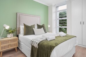 A neatly made bed with a green throw blanket, white bedding, and two rolled towels. A bedside table with a lamp and a clock is visible. - The Sands BG3 (Cape Town)