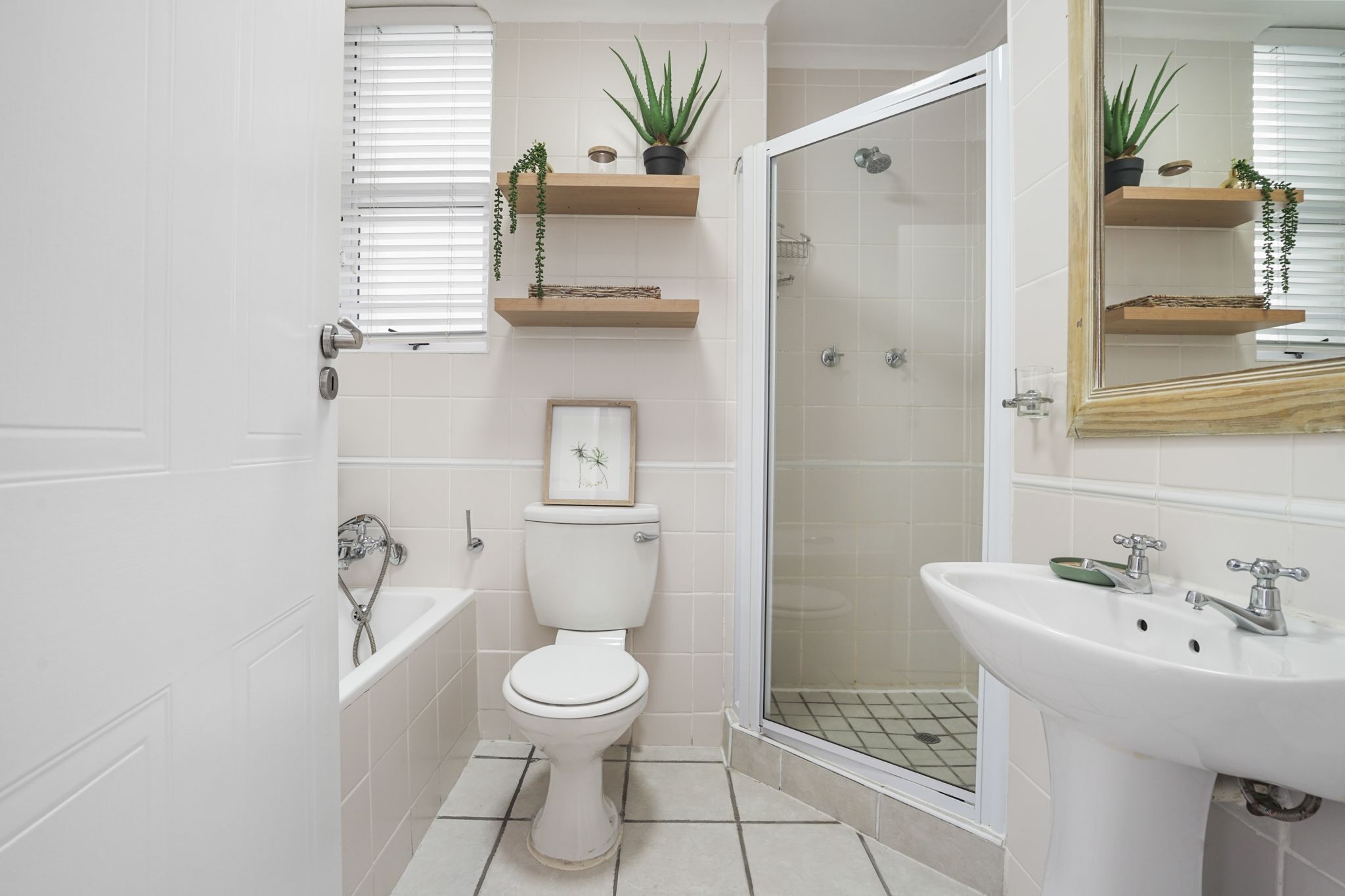 Un bagno con WC, vasca, doccia e lavandino. Scaffali in legno con piante e un quadro incorniciato sopra il WC.