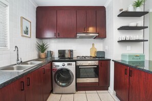 A compact kitchen with dark wooden cabinets, a stainless steel washing machine, an oven, and a toaster. - The Sands BG3 (Cape Town)