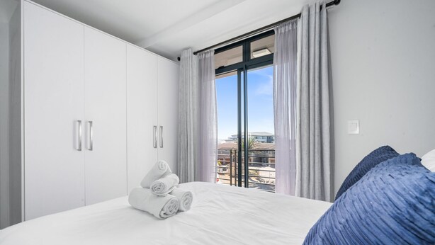 A hotel room with a bed, white curtains, and a view of buildings through the window. - Beach Break 47 (Cape Town)