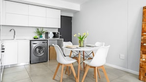 A compact kitchen with white cabinets, a washing machine, and a dining area with a table and chairs. - Beach Break 47 (Cape Town)