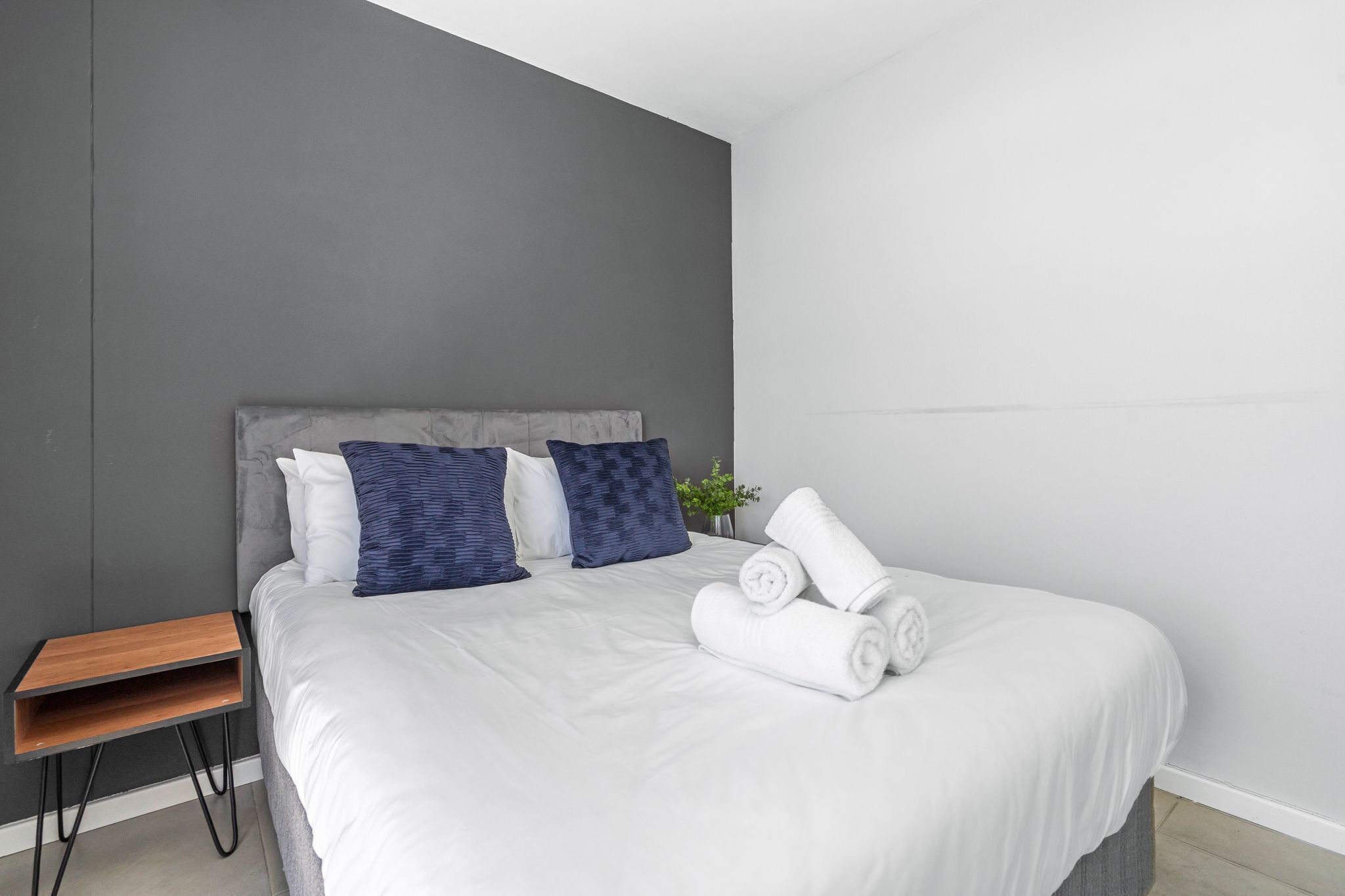 A neatly made bed with white linens and rolled towels, a grey headboard, and a small wooden nightstand with a plant.