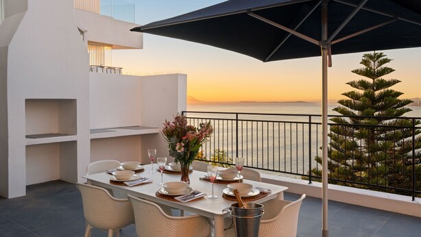A dining table set with plates, glasses, and a flower vase on a balcony overlooking the sea. - Suikerbossie 52 (Cape Town)