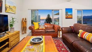 A modern living room with a brown leather sofa, a wooden coffee table, and a TV on a wooden stand. There are two large windows with a view of the city and mountains. - Suikerbossie 52 (Cape Town)