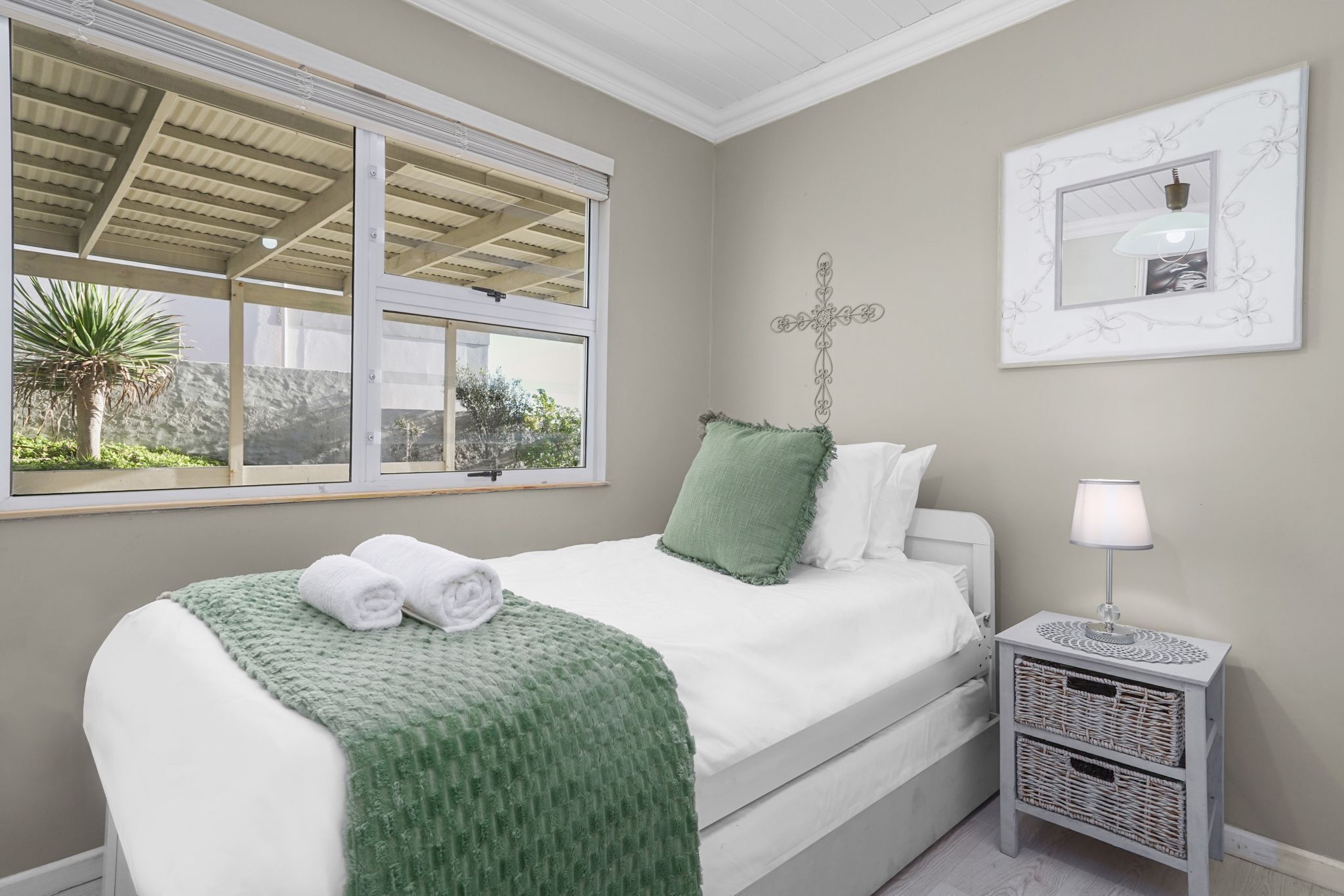 Une chambre à coucher avec un lit, une fenêtre, une croix accrochée au mur et une table de chevet avec une lampe.