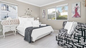 A bedroom with a large bed, a nightstand, a patterned armchair, and a window with a view of palm trees.
