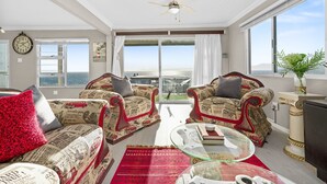 A living room with two armchairs, a coffee table, and a TV. There is a large window with a view of the sea and a balcony.