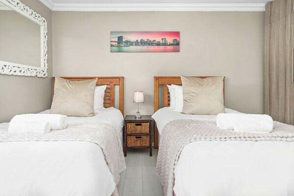 A hotel room with two beds, a nightstand with a lamp, a cityscape painting on the wall, and a large mirror. - The Tides at Gansbaai (De Kelders)