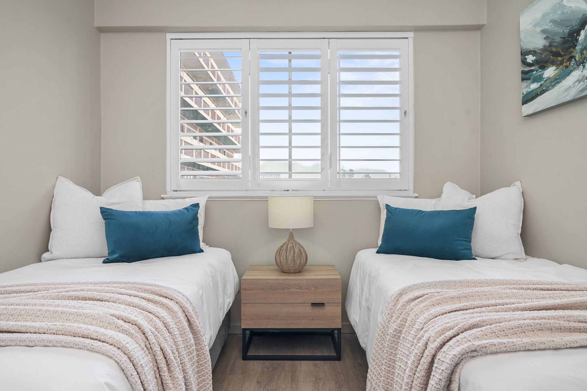 Un dormitorio con dos camas, una mesita de noche con lámpara, una ventana con persianas y un cuadro en la pared.