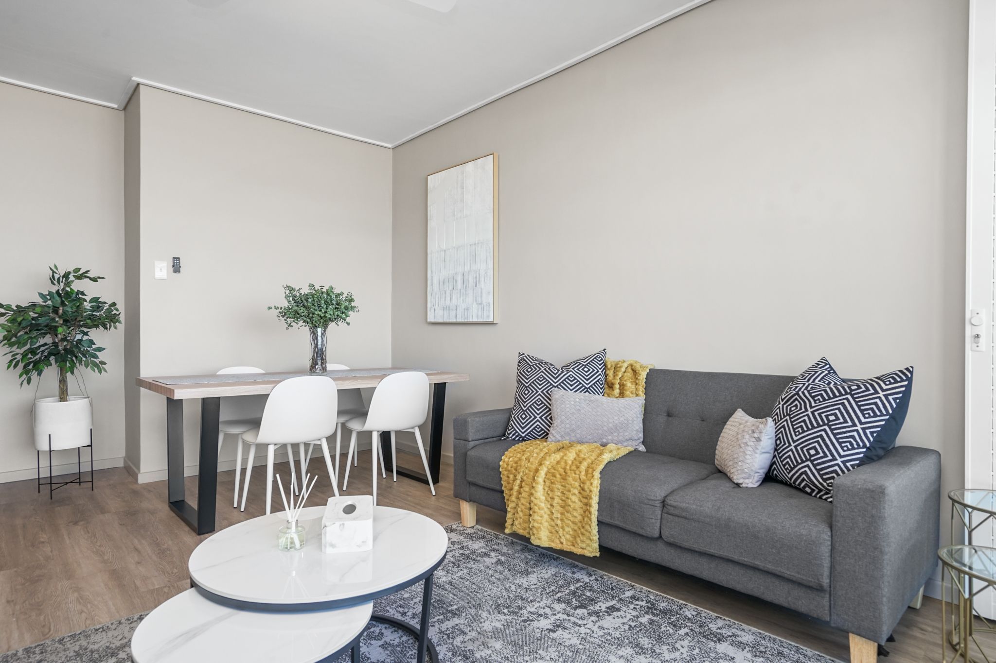 Een moderne woonkamer met een grijze bank, gestreepte kussens, een ronde salontafel en een eethoek met een witte tafel en stoelen.