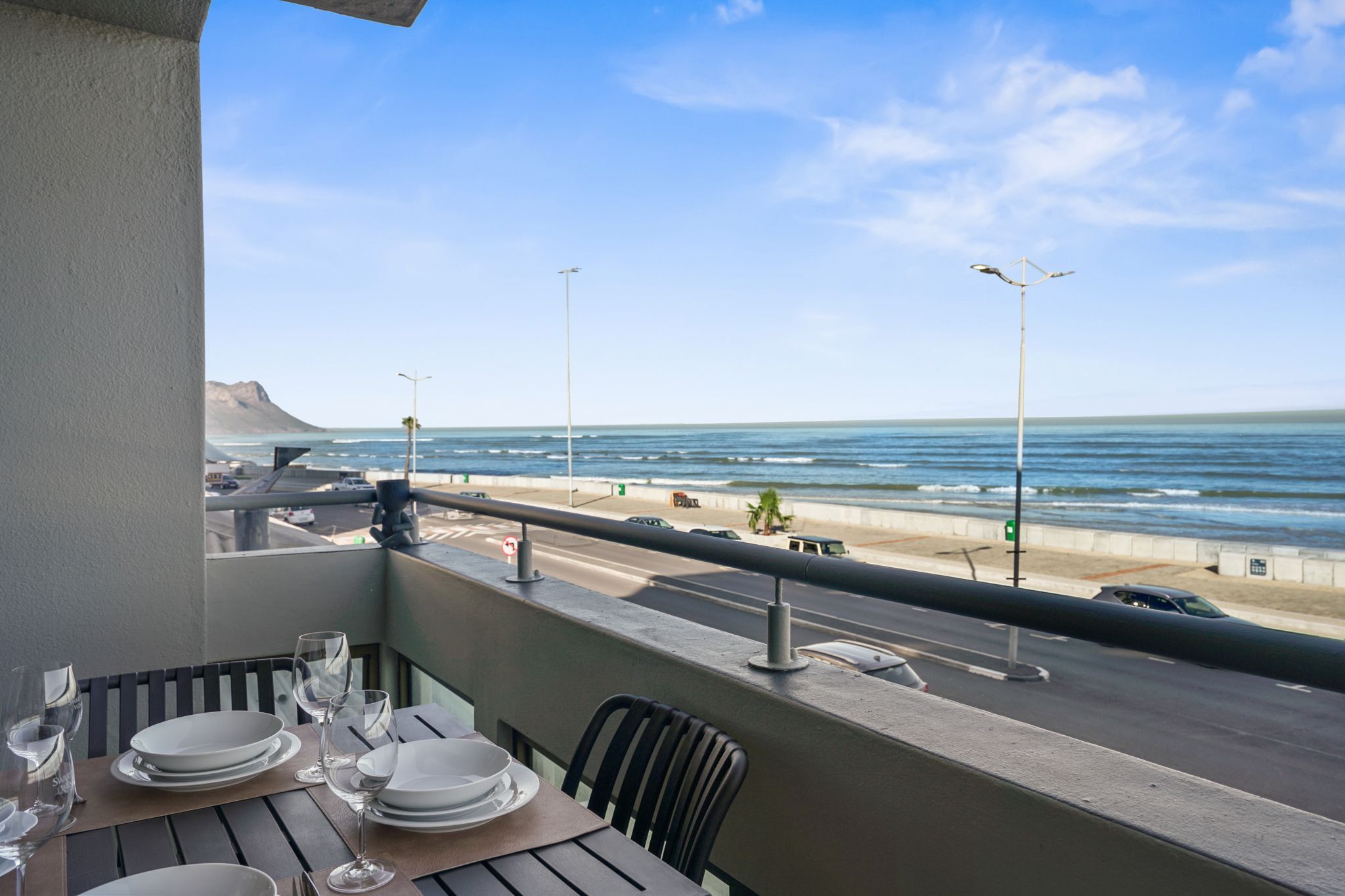 A balcony with a table set for dining, overlooking a beach with a road and vehicles, and a mountain in the distance.