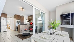 A modern kitchen with a built-in washing machine, a dining area with a white table set for four, and a sliding glass door leading to a balcony with a plant and a wine bottle. - Balmoral Heights 20 (Cape Town)