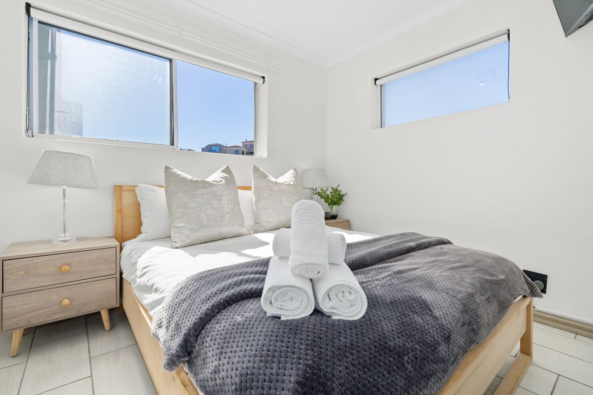 Un dormitorio con una cama, una mesita de noche, una lámpara y dos ventanas.