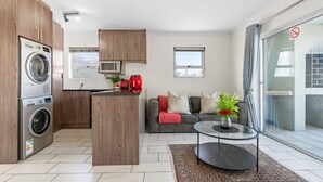 A modern kitchen with a built-in washer and dryer, a microwave, and a red kettle. There is a grey sofa with a red throw blanket and a glass coffee table with a vase of flowers. - Balmoral Heights 20 (Cape Town)