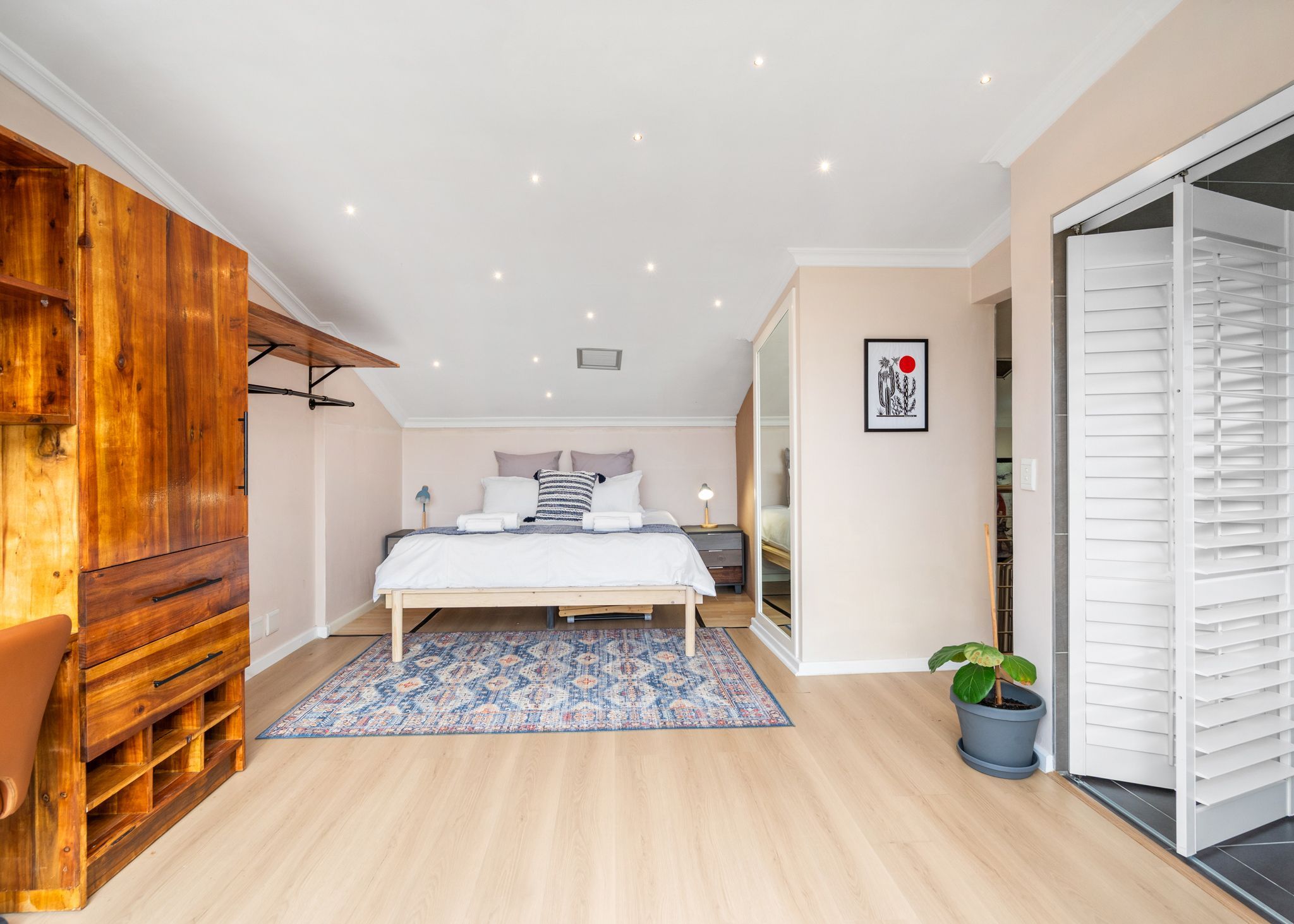 A bedroom with a bed, bedside tables, a wooden wardrobe, a mirror, a potted plant, and a sliding door.