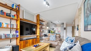 A modern living room with a large flat-screen TV, wooden bookshelves, a leather sofa, and a dining area in the background. - The Tygervalley Escape (Cape Town)