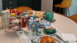 Ein Esszimmer mit gedecktem Tisch, Speisen, Getränken, einer grünen Teekanne und einer weißen Tischdecke mit Blumenmuster.