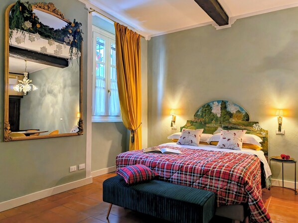 Ein Schlafzimmer mit Bett, Bank, großem Spiegel, Fenster mit Vorhängen und Wandleuchten.
