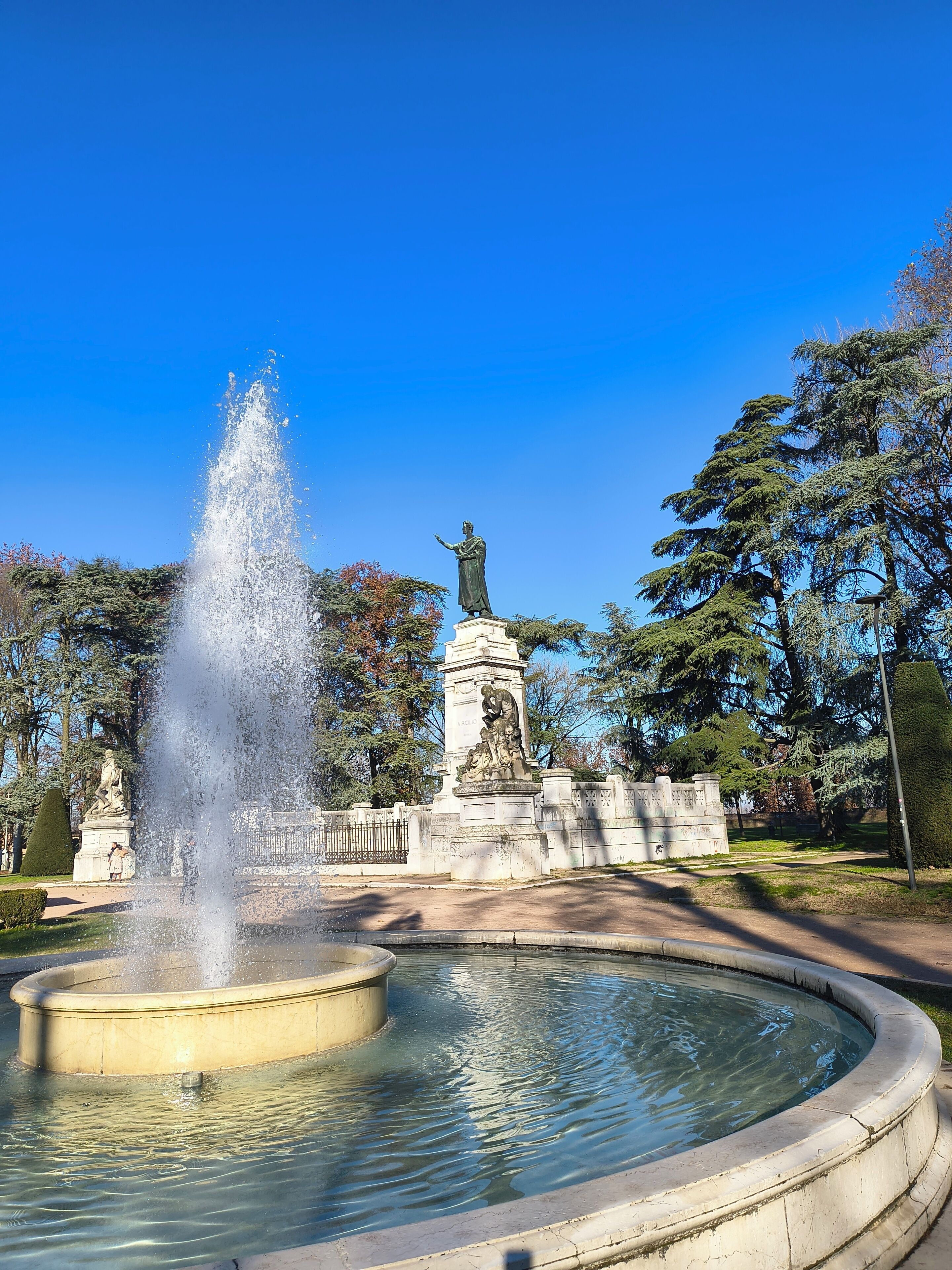 Una fontana con una statua al centro, circondata da alberi e un cielo sereno.