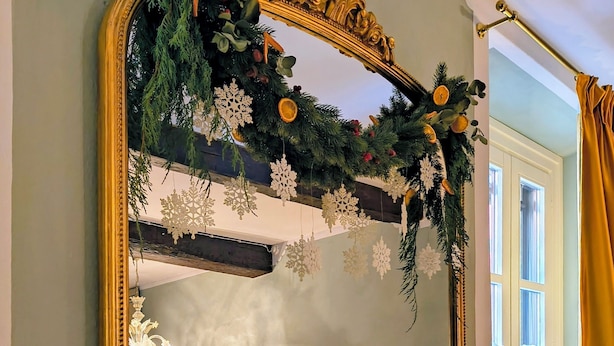 Ein Schlafzimmer mit einem großen, verzierten Spiegel, einem Bett mit karierter Bettwäsche, einem kleinen Tisch mit Stuhl und einem Fenster mit Vorhängen.