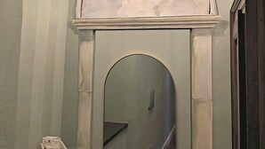 Ein Badezimmer mit gewölbter Decke, einer Wandleuchte, einem Spiegel, einer Toilette und einem kleinen Regal mit einem Korb.