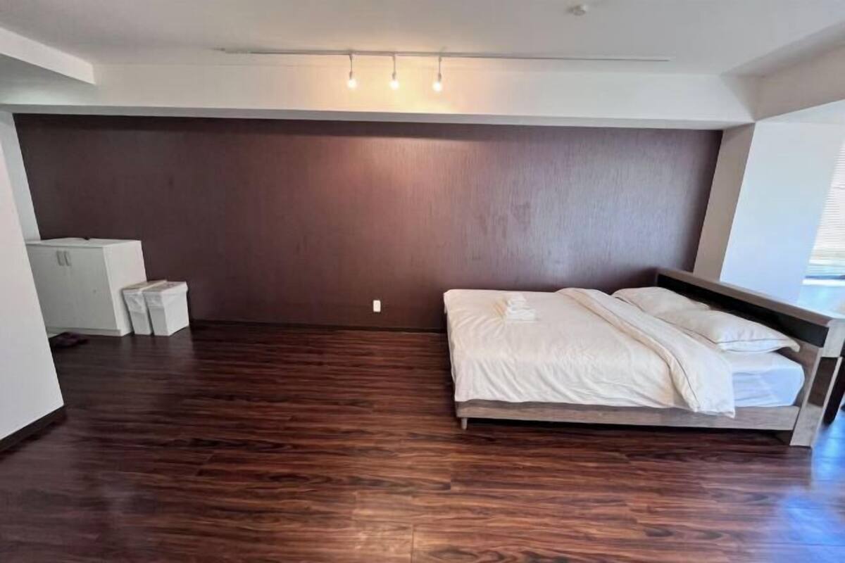 A modern hotel room with a wooden floor, a bed with white linens, a built-in bench, and a wall-mounted light fixture.