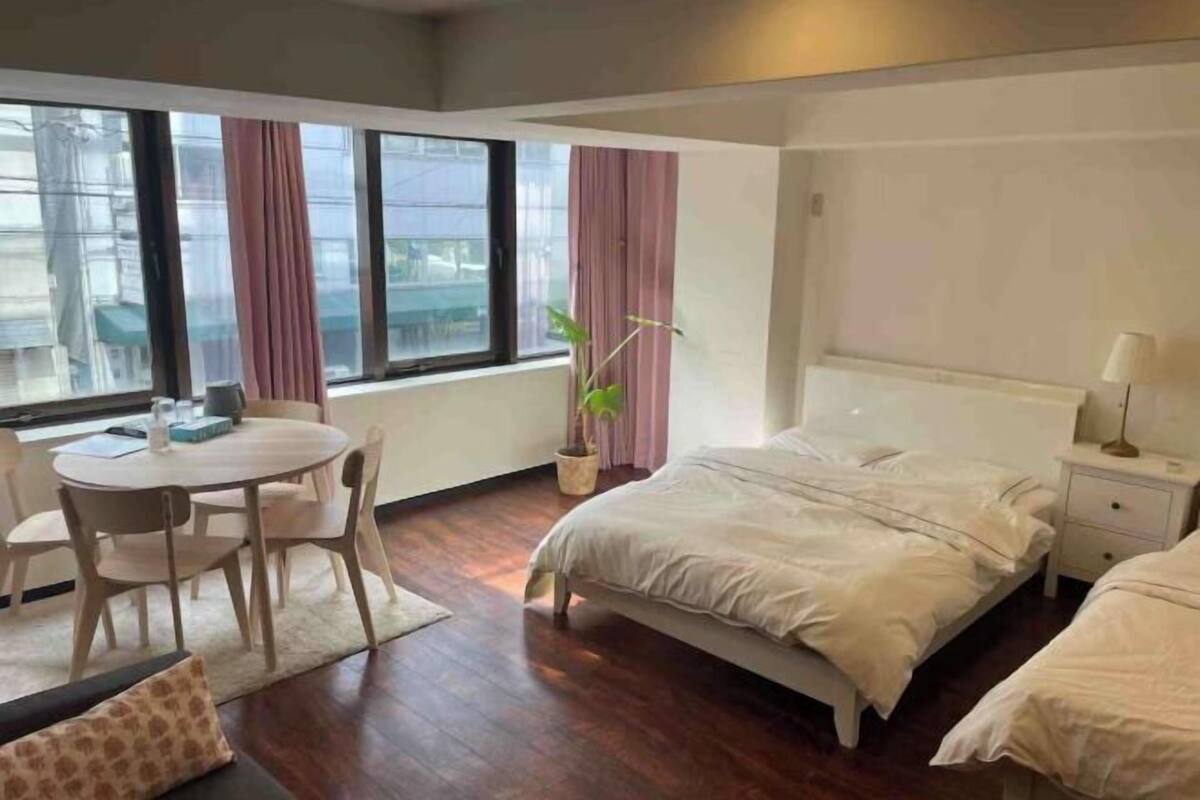 A hotel room with a bed, a small table, a chair, a potted plant, and a window with curtains.