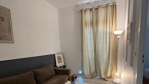 Uma sala de estar com um sofá marrom, uma mesa de centro branca, um vaso com flores, um abajur e uma grande janela com cortinas.
