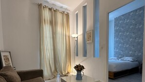 Uma sala de estar moderna com um sofá marrom, uma mesa de centro branca e um vaso azul com flores secas. Uma cama com cabeceira estampada pode ser vista ao fundo.
