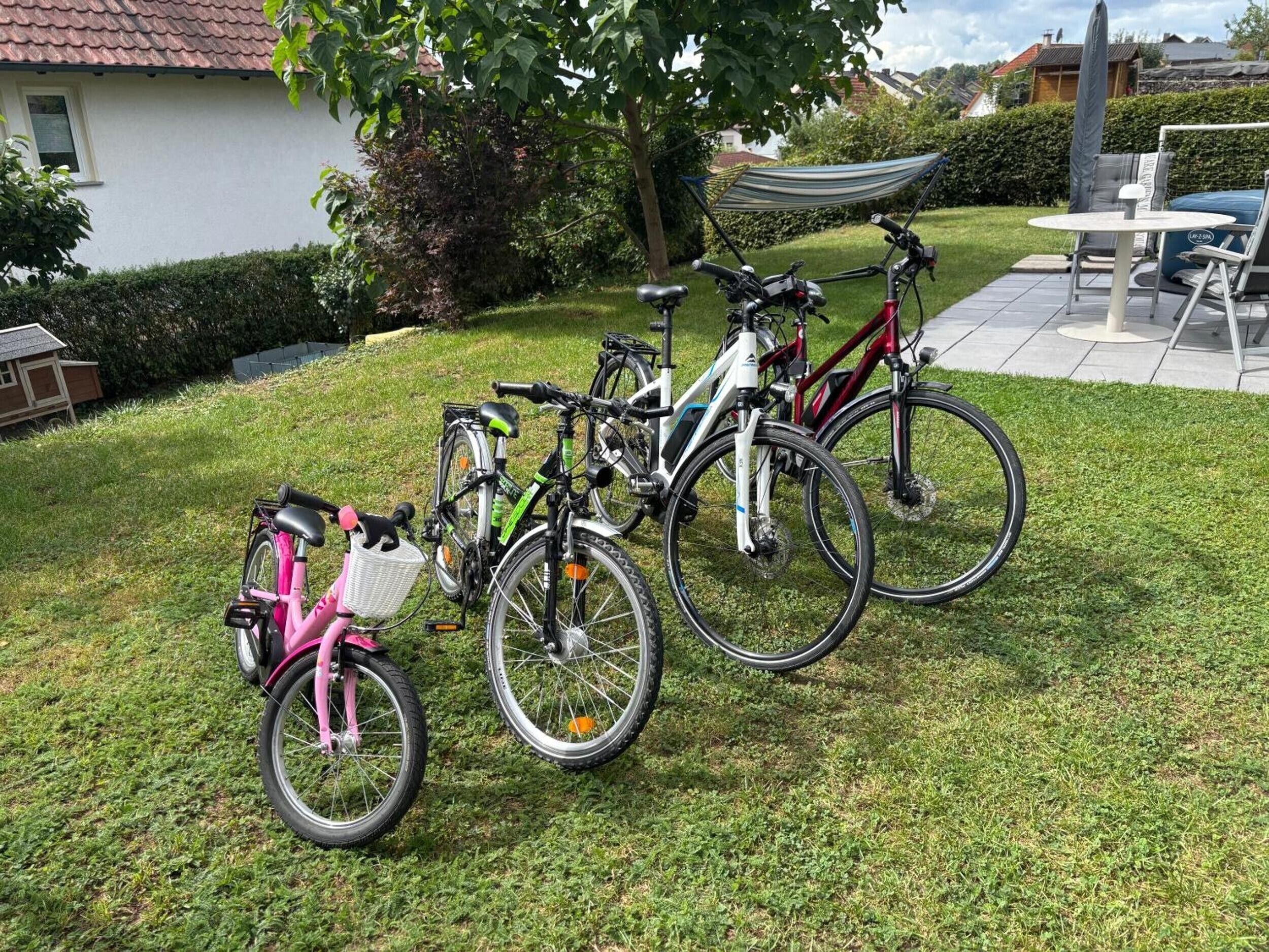Des vélos sont garés sur une pelouse verdoyante, un bâtiment blanc se dresse à proximité, et un hamac est visible au fond.