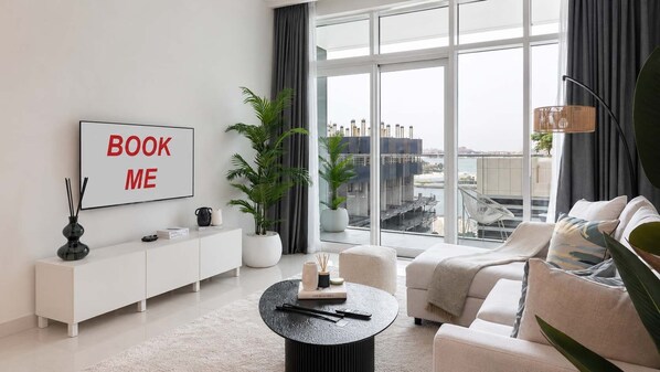 A modern living room with a sofa, a coffee table, a bookshelf, and a large window with a city view.