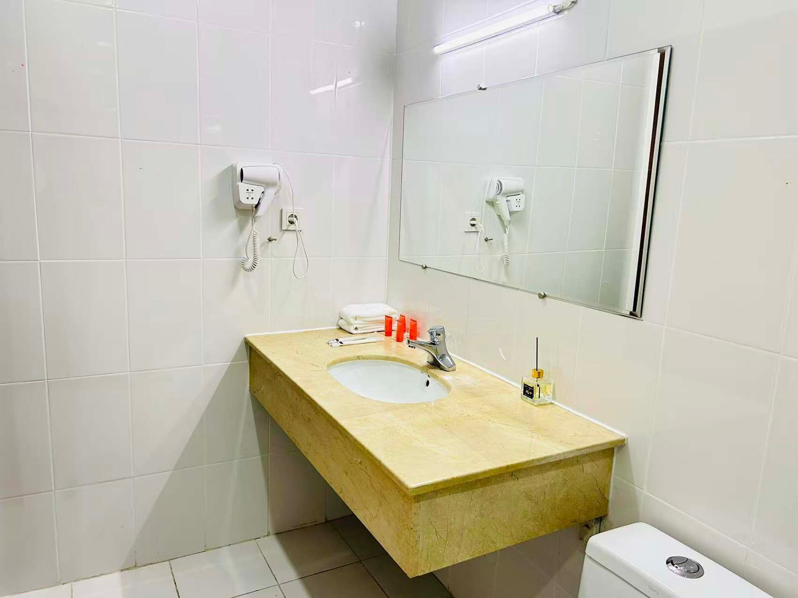Ein Badezimmer mit einem beigefarbenem Waschbecken, weißen Fliesen, einem Spiegel, einer weißen Toilette und einem an der Wand befestigten Föhn.