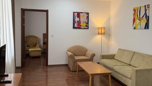 A living room with a wooden floor, a sofa, a chair, a coffee table, and a TV. There is a door leading to another room. - Fude Hotel (Tbilisi)