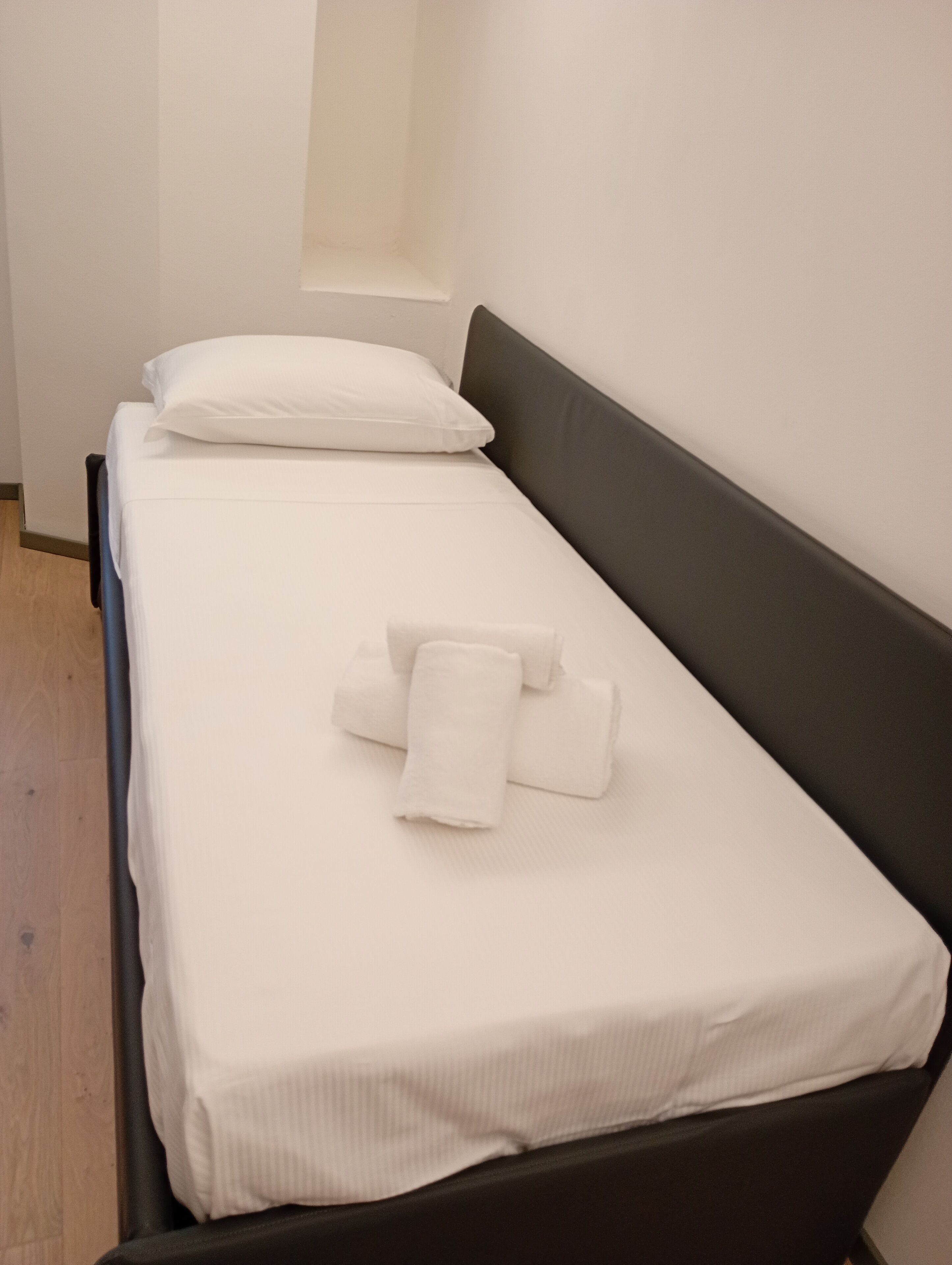 A single bed with white bedding and pillows, a dark headboard, and a wooden floor.