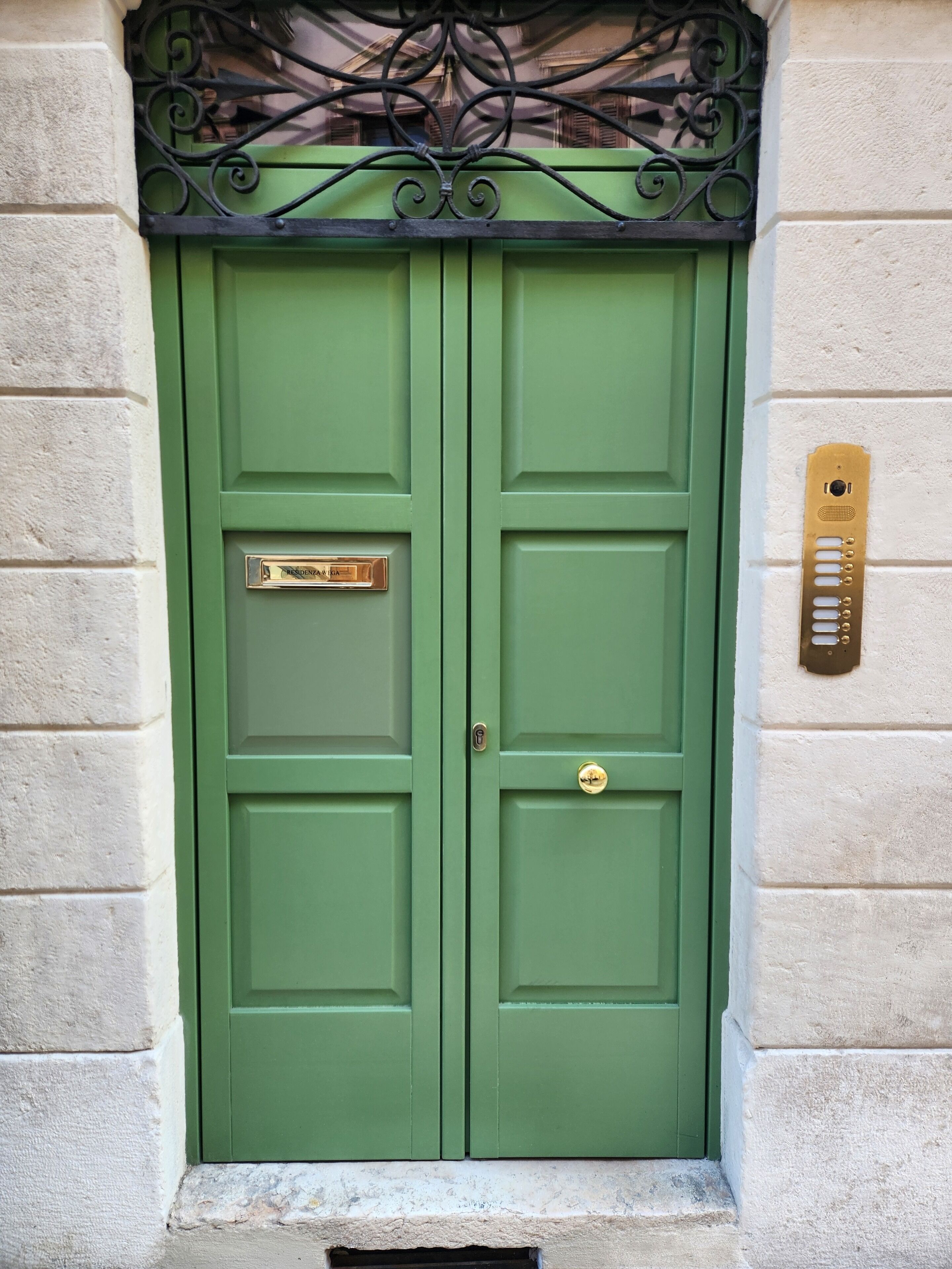 A green double door with a brass mail slot and a brass intercom on a textured wall.