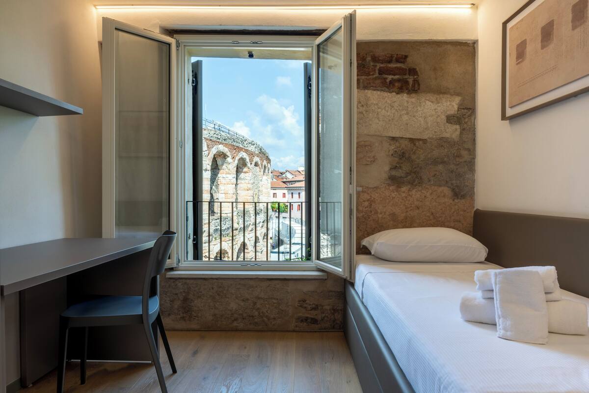 A bedroom with a bed, a desk, and a window with a view of a historic building.