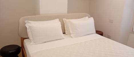 Una cama bien tendida con ropa de cama blanca y dos almohadas, una mesita de noche de madera con un objeto negro encima, y una pared blanca lisa con un techo texturizado.