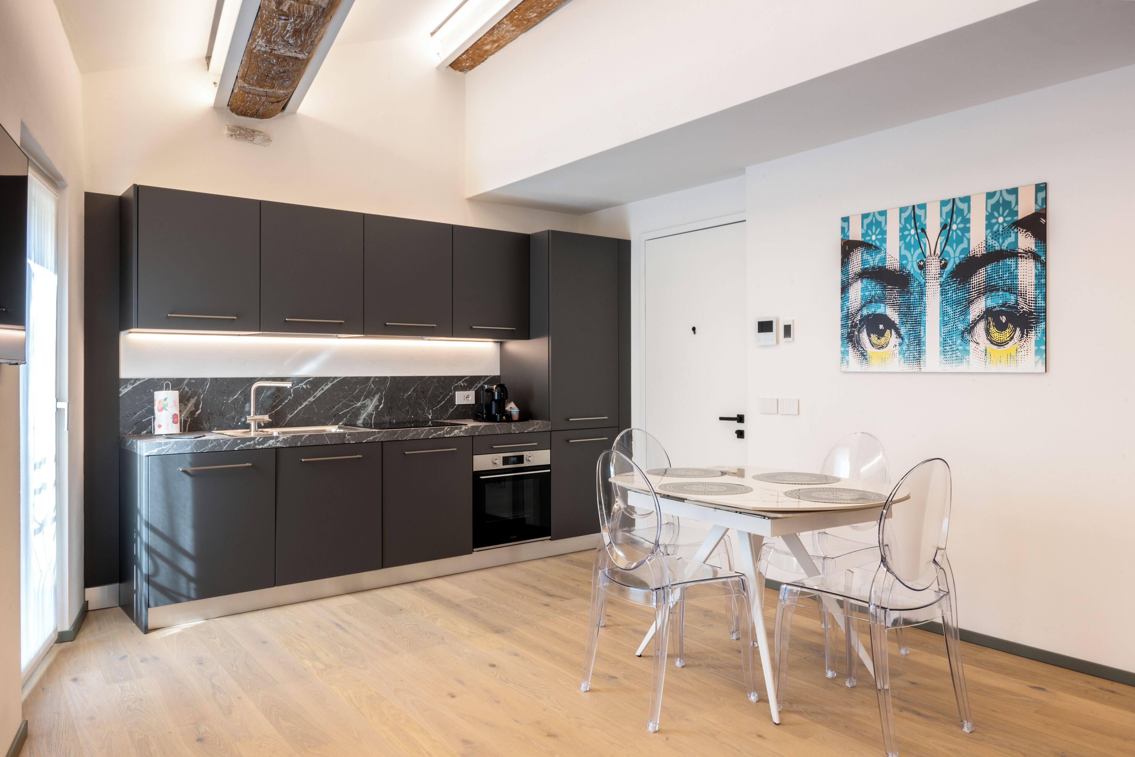 Une cuisine moderne avec des meubles de couleur sombre, un plan de travail en marbre et un coin repas comprenant une table et des chaises en verre transparent.