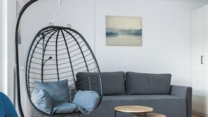 Un salon moderne avec un canapé gris, un fauteuil suspendu avec des coussins, une table ronde en bois et une petite table d’appoint.
