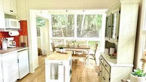 Une cuisine avec un coin repas, un plancher en bois, des armoires blanches, un four à micro-ondes et une grande fenêtre donnant sur la forêt.